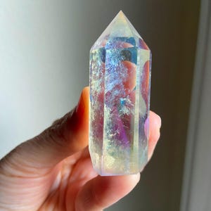 Puede incluir: Una punta de cristal hexagonal transparente con un revestimiento iridiscente similar a un arco&iacute;ris. La piedra pulida presenta una parte superior puntiaguda y facetas verticales que reflejan la luz, mostrando tonos cambiantes de azul, morado y oro.