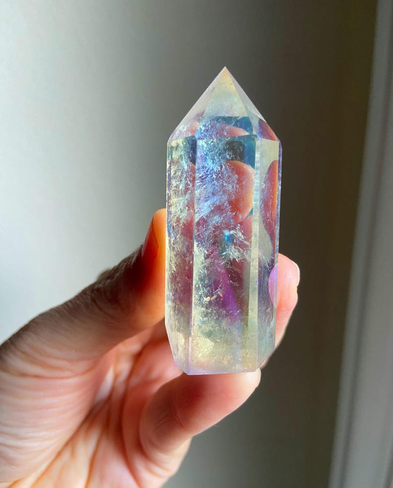 Puede incluir: Una punta de cristal hexagonal transparente con un revestimiento iridiscente similar a un arco&iacute;ris. La piedra pulida presenta una parte superior puntiaguda y facetas verticales que reflejan la luz, mostrando tonos cambiantes de azul, morado y oro.