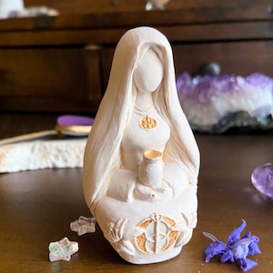 May include: White ceramic figurine of a woman with long hair, wearing a robe and holding a chalice. The figurine has gold accents and is sitting on a base with a circular design.