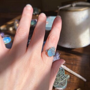 May include: A silver ring featuring a raw, light blue gemstone set in a prong setting. The stone has an irregular, jagged shape with a translucent appearance. The metal band is thin and polished, contrasting with the textured surface of the stone.