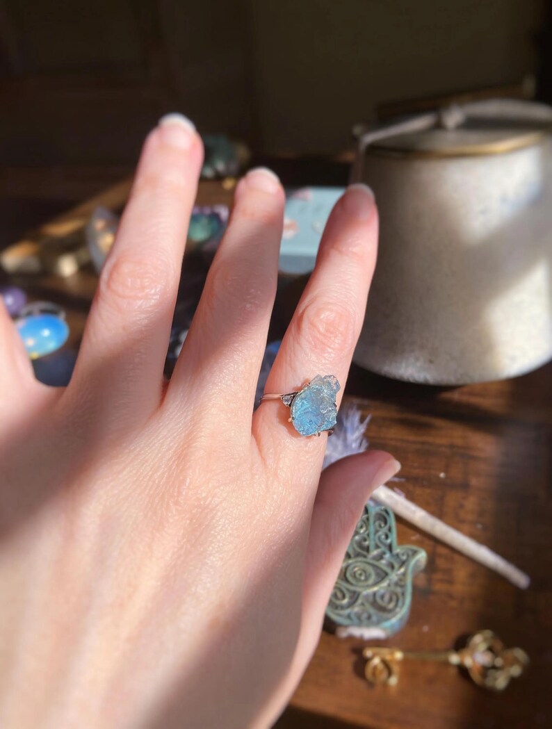 May include: A silver ring featuring a raw, light blue gemstone set in a prong setting. The stone has an irregular, jagged shape with a translucent appearance. The metal band is thin and polished, contrasting with the textured surface of the stone.