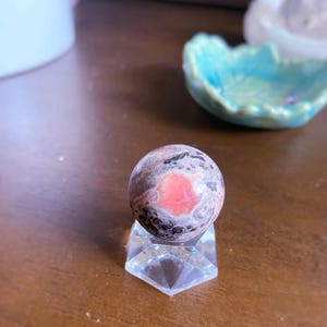 May include: A spherical gemstone with pink and gray patterns, displayed on a clear, faceted crystal stand. The stone has a prominent pink center and is approximately 1 inch in diameter. The stand is hexagonal and reflects light.