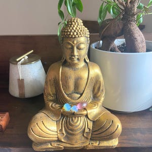May include: A gold Buddha statue sitting in meditation with three small, colorful stones in its hands. The statue is on a wooden surface with a pink stone and a white ceramic container in the background.