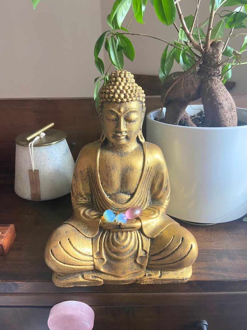 May include: A gold Buddha statue sitting in meditation with three small, colorful stones in its hands. The statue is on a wooden surface with a pink stone and a white ceramic container in the background.