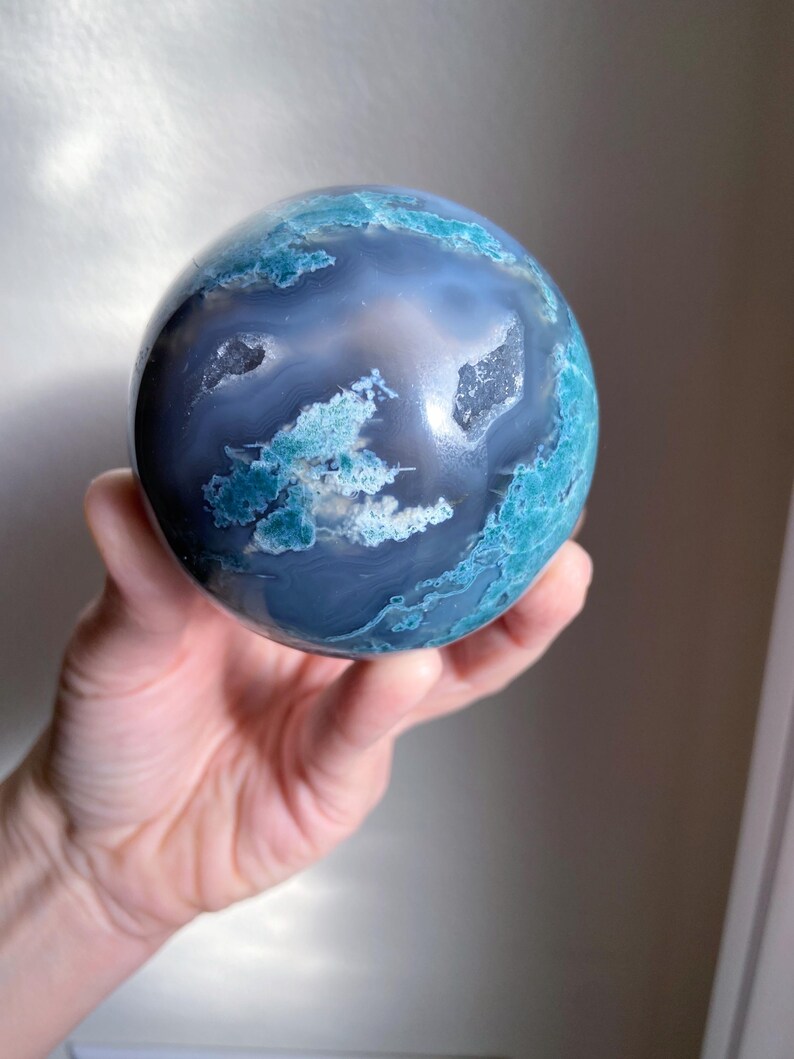 Pu&ograve; includere: Una gemma sferica lucidata con un motivo a spirale di blu e bianco, che ricorda un pianeta in miniatura. La pietra presenta aree di formazioni cristalline ed &egrave; tenuta in una mano.