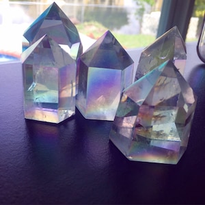 May include: Five iridescent quartz crystal points with a prism effect. The crystals are clear with a rainbow sheen, reflecting colors of blue, purple, and yellow. They are arranged on a dark surface, with a window in the background.