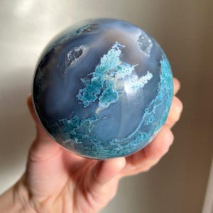 Pu&ograve; includere: Una sfera di agata lucidata, che mostra una miscela di tonalit&agrave; blu e verde acqua. La pietra presenta motivi intricati e formazioni cristalline, creando un oggetto decorativo unico. La sfera &egrave; tenuta in una mano.