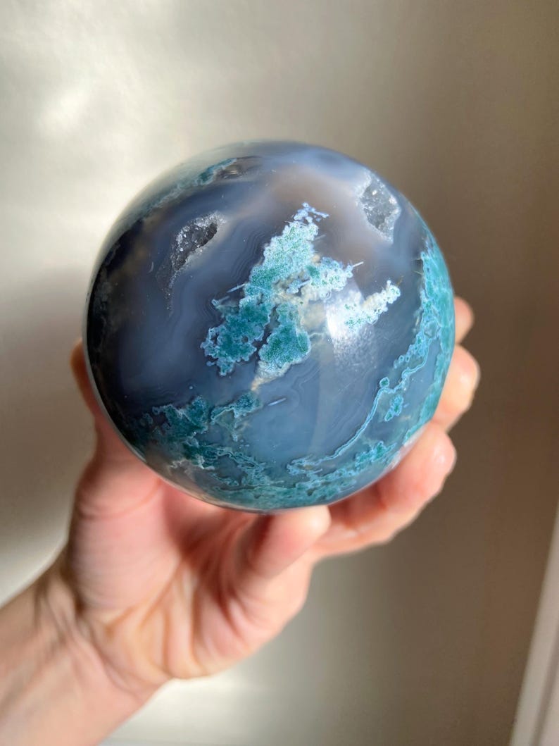 Pu&ograve; includere: Una sfera di agata lucidata, che mostra una miscela di tonalit&agrave; blu e verde acqua. La pietra presenta motivi intricati e formazioni cristalline, creando un oggetto decorativo unico. La sfera &egrave; tenuta in una mano.