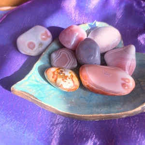 May include: A collection of polished stones in various colors, including pink, white, and orange, displayed on a turquoise leaf-shaped dish. The stones have unique patterns and textures. The dish sits on a purple fabric surface.