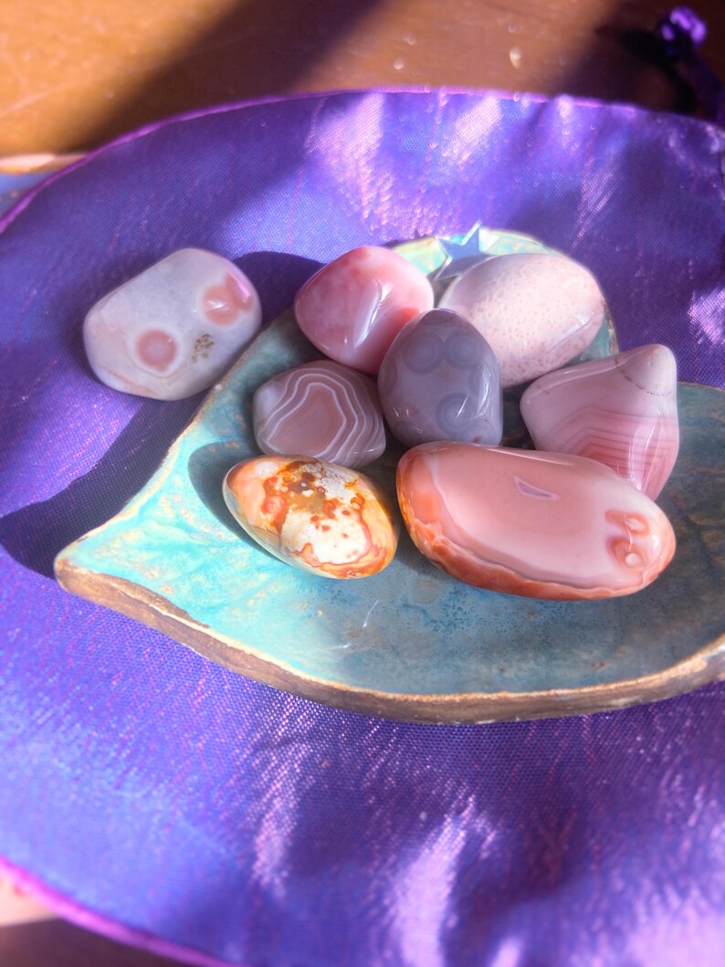 May include: A collection of polished stones in various colors, including pink, white, and orange, displayed on a turquoise leaf-shaped dish. The stones have unique patterns and textures. The dish sits on a purple fabric surface.
