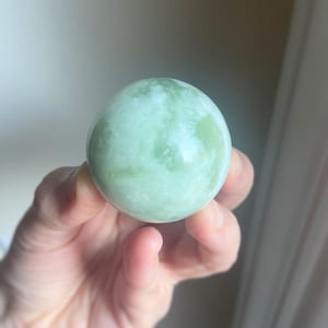 May include: A light green, polished stone sphere with white marbling. The sphere is held in a hand, showcasing its smooth, rounded form. The stone's color is a soft, muted green, with subtle variations in tone.