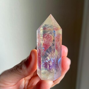 Puede incluir: Una punta de cristal transparente de seis lados con un revestimiento met&aacute;lico iridiscente similar al arco&iacute;ris. La piedra presenta fracturas e inclusiones internas que captan la luz, mostrando destellos de tonos rosa, morado y dorado.