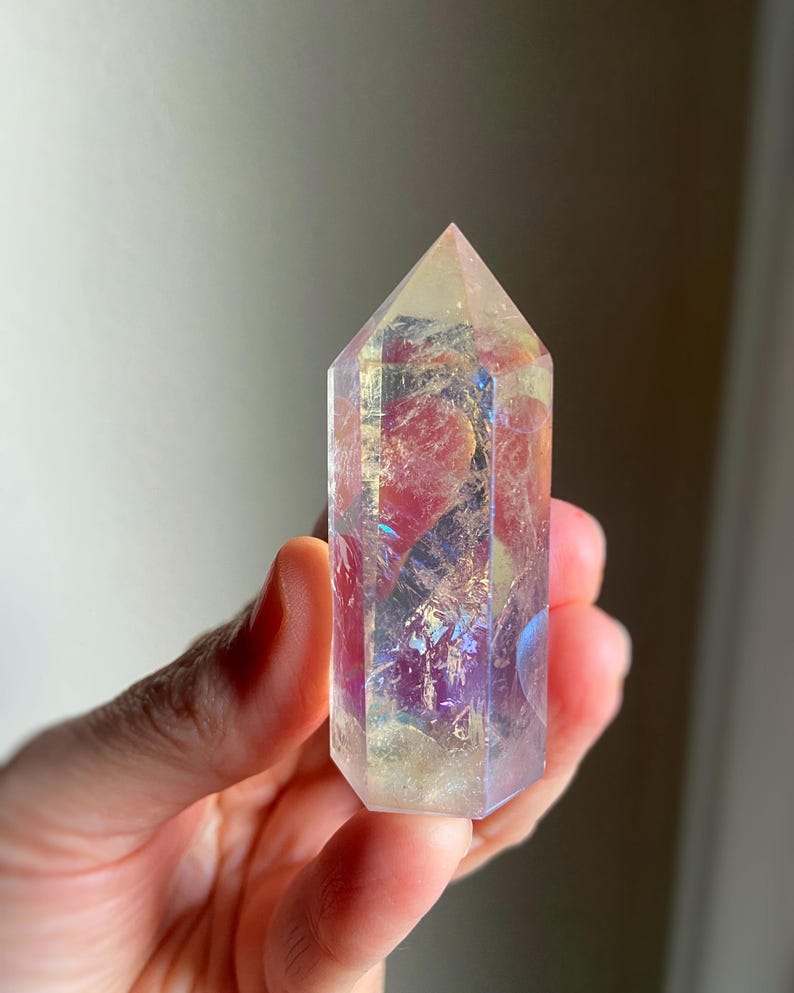 Puede incluir: Una punta de cristal transparente de seis lados con un revestimiento met&aacute;lico iridiscente similar al arco&iacute;ris. La piedra presenta fracturas e inclusiones internas que captan la luz, mostrando destellos de tonos rosa, morado y dorado.