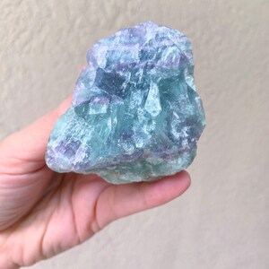 May include: A hand holds a raw, natural fluorite crystal. The stone displays a mix of blue, green, and purple hues. The crystal's surface is rough and textured, with visible crystalline structures. The background is a neutral, light beige color.