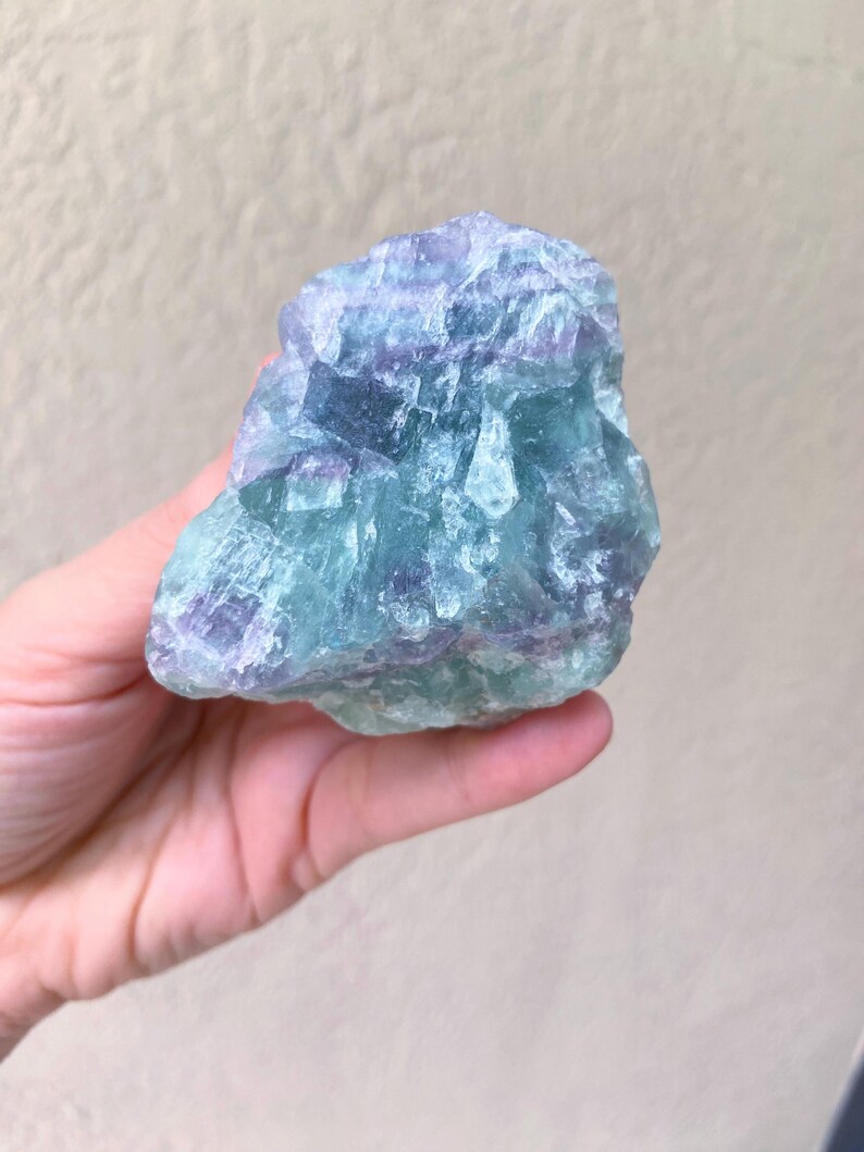May include: A hand holds a raw, natural fluorite crystal. The stone displays a mix of blue, green, and purple hues. The crystal's surface is rough and textured, with visible crystalline structures. The background is a neutral, light beige color.