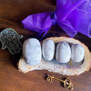 May include: Four white and gray Howlite stones are displayed on a decorative wooden slice, with a gold-colored key and a silver Hamsa hand. A purple ribbon adds a touch of color. The text "One With Gems" is visible.