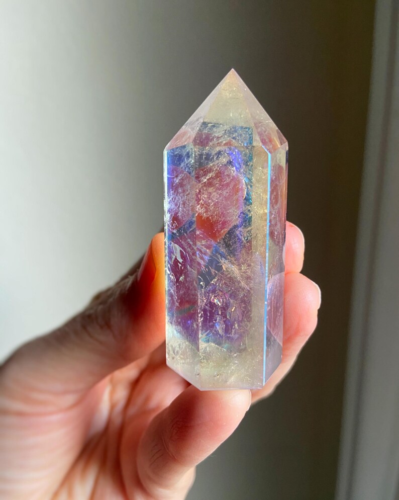 Puede incluir: Una punta de cristal hexagonal con acabado pulido, que presenta una base transl&uacute;cida amarilla y clara con un revestimiento met&aacute;lico iridiscente tipo arco&iacute;ris que refleja tonos azules, p&uacute;rpuras y dorados sobre su superficie facetada.