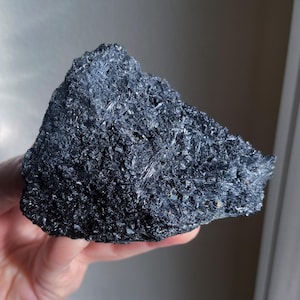 May include: A dark, irregularly shaped mineral specimen with a rough, crystalline surface. The color is a deep, lustrous black, with visible reflective facets. The overall texture appears coarse and jagged, suggesting a natural, unpolished state. The mineral is held in a hand.