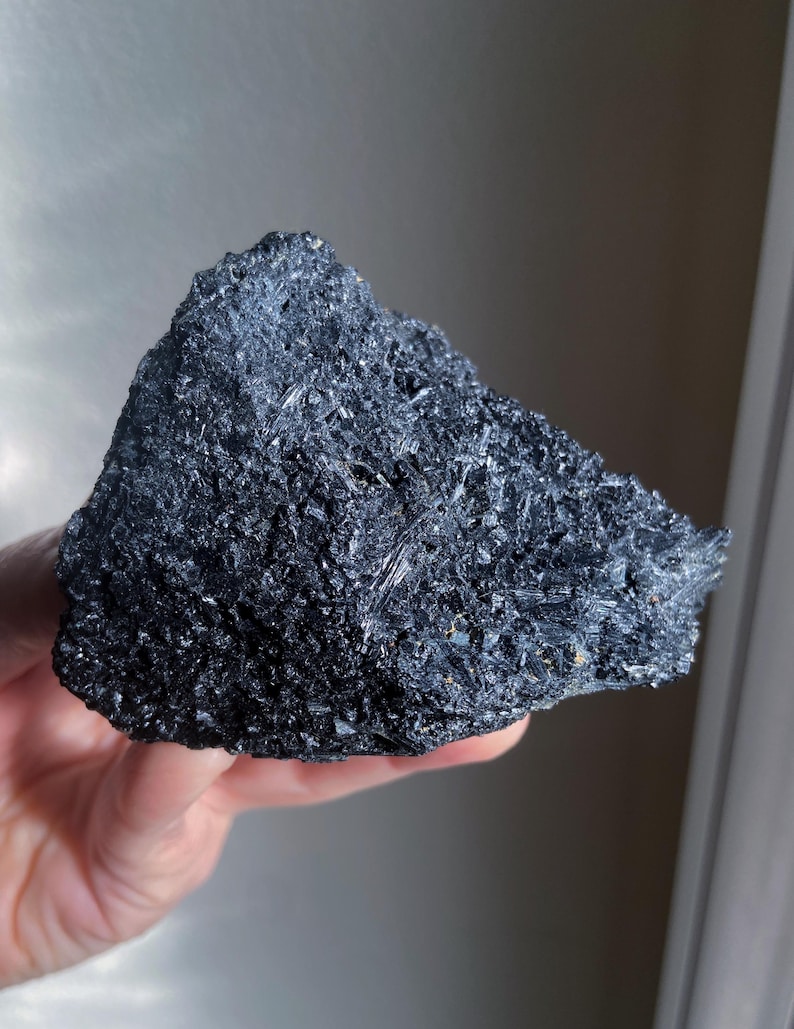 May include: A dark, irregularly shaped mineral specimen with a rough, crystalline surface. The color is a deep, lustrous black, with visible reflective facets. The overall texture appears coarse and jagged, suggesting a natural, unpolished state. The mineral is held in a hand.