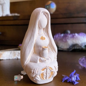 May include: White ceramic figurine of a woman with long hair, wearing a robe and holding a small cup. The figurine has gold accents and is sitting in a meditative pose.