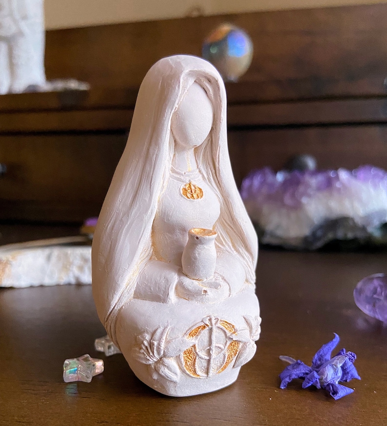 May include: White ceramic figurine of a woman with long hair, wearing a robe and holding a small cup. The figurine has gold accents and is sitting in a meditative pose.