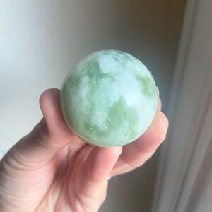 May include: A light green sphere with white marbling is held in a hand. The sphere has a smooth, polished surface and a marbled pattern of light and dark green hues. The object is a decorative item or a natural stone.