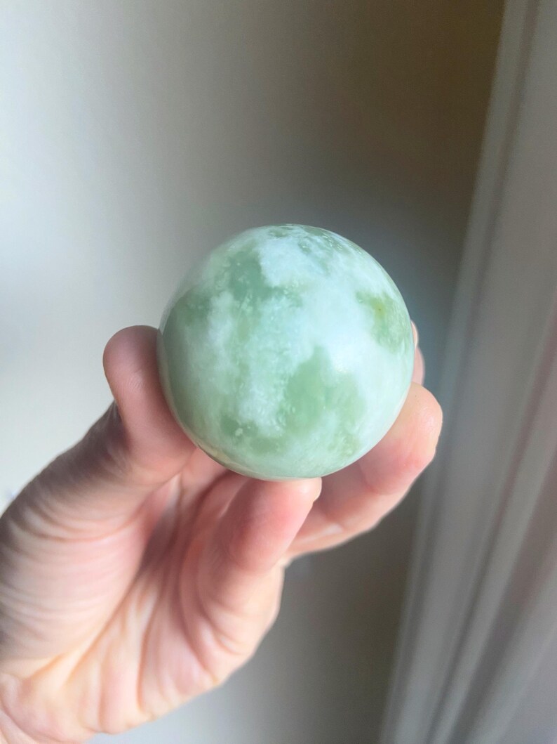 May include: A light green sphere with white marbling is held in a hand. The sphere has a smooth, polished surface and a marbled pattern of light and dark green hues. The object is a decorative item or a natural stone.