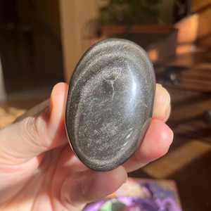 May include: A polished, oval-shaped obsidian stone with silver inclusions. The stone is a dark gray color with a glossy finish. The inclusions create a shimmering effect. The stone is held in a hand, suggesting its size and portability. This is a gemstone.