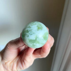 May include: A round, polished stone sphere with a mottled green and white pattern. The stone is held in a hand, showcasing its smooth surface and natural color variations. The sphere's size is approximately 2 inches in diameter.