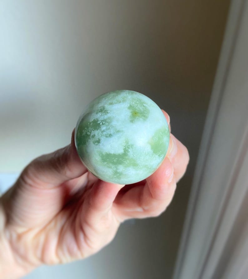 May include: A round, polished stone sphere with a mottled green and white pattern. The stone is held in a hand, showcasing its smooth surface and natural color variations. The sphere's size is approximately 2 inches in diameter.
