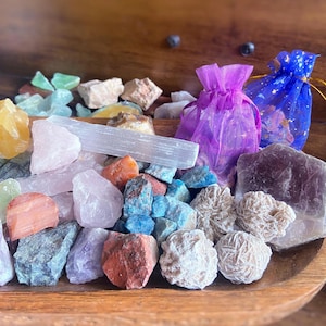 May include: A wooden bowl filled with a variety of colorful crystals and gemstones. The collection includes rose quartz, selenite, blue kyanite, and desert roses. Two small drawstring bags, one purple and one blue, are also present.