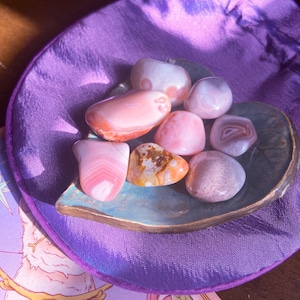 May include: A collection of polished gemstones in shades of pink, peach, and brown, displayed on a leaf-shaped ceramic dish. The dish sits on a purple satin pouch. The gemstones vary in size and pattern, with some showing banded layers.