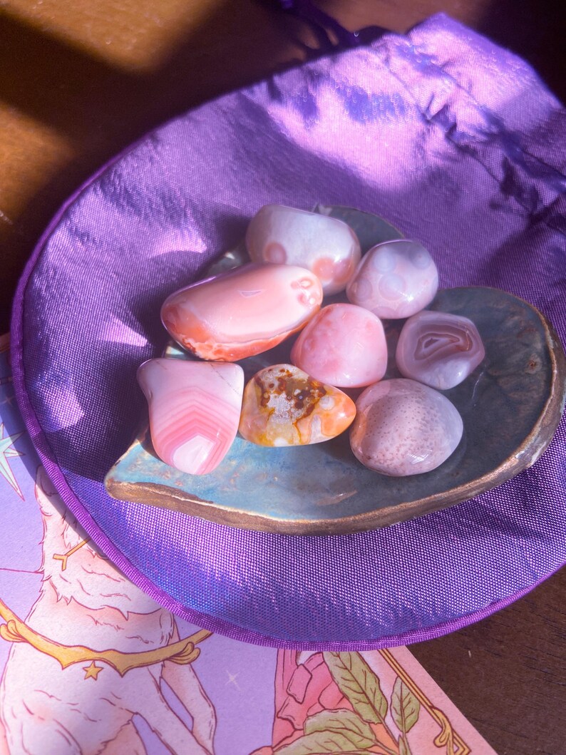 May include: A collection of polished gemstones in shades of pink, peach, and brown, displayed on a leaf-shaped ceramic dish. The dish sits on a purple satin pouch. The gemstones vary in size and pattern, with some showing banded layers.