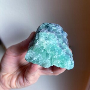 May include: A raw, natural fluorite crystal with a mix of blue and green hues. The stone has a rough, textured surface and is held in a hand. The crystal's colors blend together, creating a unique and eye-catching appearance.