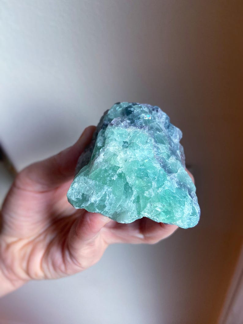 May include: A raw, natural fluorite crystal with a mix of blue and green hues. The stone has a rough, textured surface and is held in a hand. The crystal's colors blend together, creating a unique and eye-catching appearance.