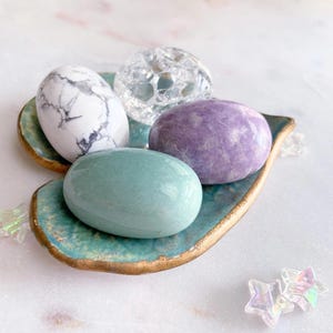 May include: A collection of polished stones in various colors, including white with gray veining, clear, purple, and teal, arranged on a teal and gold leaf-shaped dish. Decorative star-shaped crystals are scattered nearby.