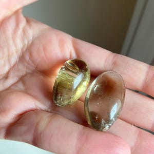 May include: Two smooth, oval-shaped polished gemstones. One stone is a translucent golden-yellow color with visible internal inclusions, while the other is a translucent brownish-grey color with a clear, glassy surface and subtle internal texture.