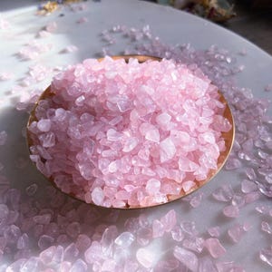 May include: A small bowl filled with pink rose quartz chips. The bowl is made of gold metal and is sitting on a white surface. There are more rose quartz chips scattered around the bowl.