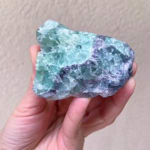 May include: A hand holding a raw fluorite crystal. The crystal displays a mix of green, blue, and purple hues. The rough texture and natural form of the stone are visible, showcasing its mineral composition. The background is a neutral beige.