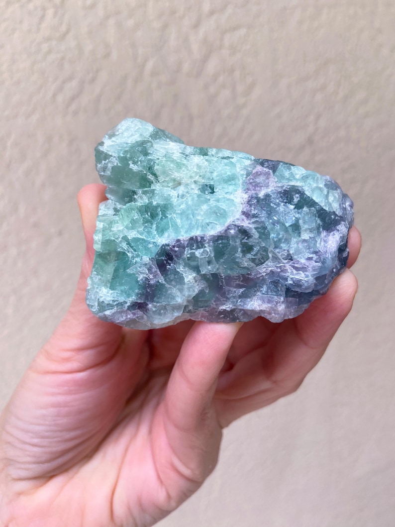 May include: A hand holding a raw fluorite crystal. The crystal displays a mix of green, blue, and purple hues. The rough texture and natural form of the stone are visible, showcasing its mineral composition. The background is a neutral beige.