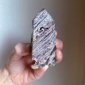 May include: A natural, multi-colored crystal point. The stone features layers of white, purple, and reddish-brown hues. The crystal has a textured surface and a pointed top. The crystal is held in a hand.