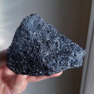 May include: A dark, irregularly shaped mineral specimen with a rough, crystalline surface. The color is a deep, lustrous black, with visible reflective facets. The overall texture appears coarse and jagged, suggesting a natural, unpolished state. The mineral is held in a hand.