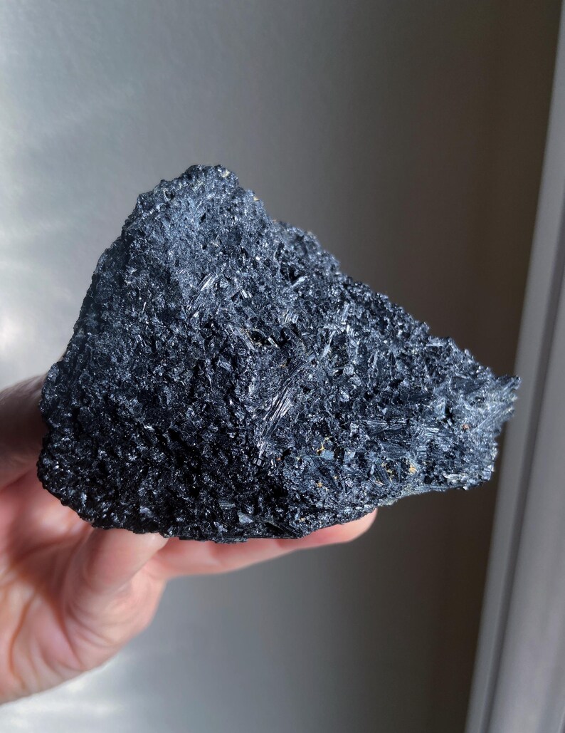 May include: A dark, irregularly shaped mineral specimen with a rough, crystalline surface. The color is a deep, lustrous black, with visible reflective facets. The overall texture appears coarse and jagged, suggesting a natural, unpolished state. The mineral is held in a hand.