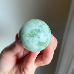 May include: A smooth, spherical, green and white stone sphere. The stone has a marbled appearance with varying shades of green and white. The sphere is held in a hand, showcasing its size and texture. This decorative item could be used as a paperweight or a decorative accent.