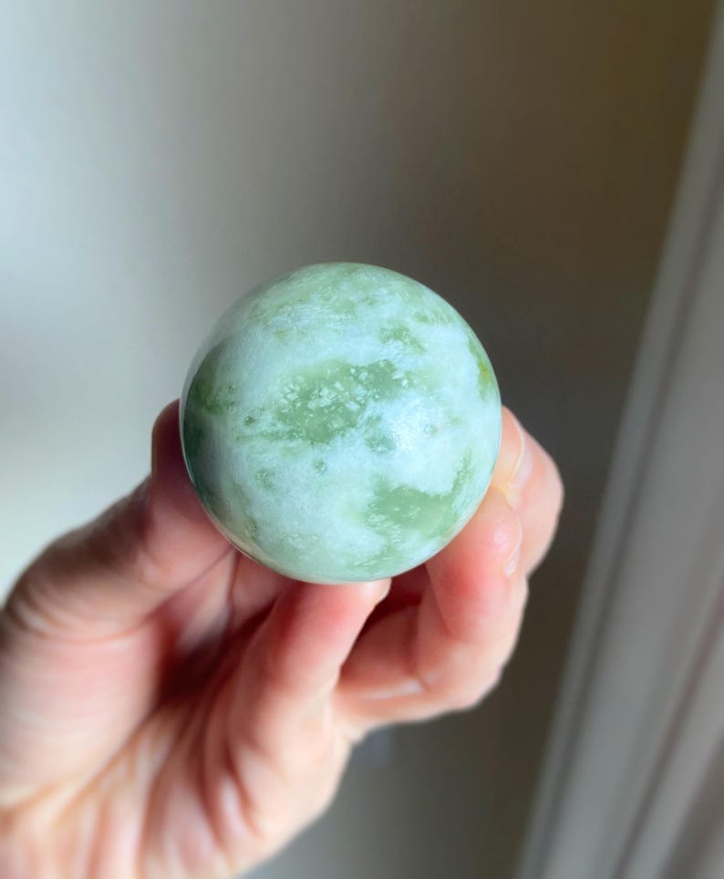 May include: A smooth, spherical, green and white stone sphere. The stone has a marbled appearance with varying shades of green and white. The sphere is held in a hand, showcasing its size and texture. This decorative item could be used as a paperweight or a decorative accent.