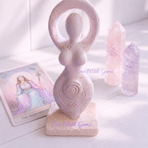 May include: A pale lavender goddess figurine with a circular head, raised arms, and a spiral design on the abdomen, featuring intricate floral carvings. The statue stands on a square stone base, accompanied by a tarot card and two crystal points.