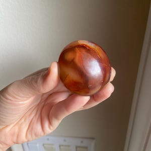 May include: A polished, spherical carnelian gemstone displaying a vibrant range of deep orange, reddish-brown, and translucent amber hues with natural internal inclusions and a smooth, glossy surface texture that reflects light across its rounded form.