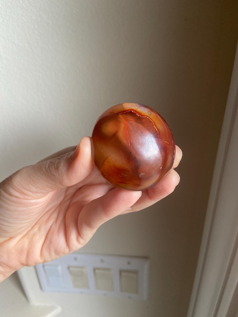 May include: A polished, spherical carnelian gemstone displaying a vibrant range of deep orange, reddish-brown, and translucent amber hues with natural internal inclusions and a smooth, glossy surface texture that reflects light across its rounded form.