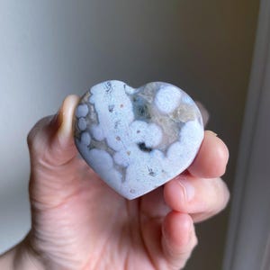 May include: A heart-shaped stone with a unique pattern of white, gray, and black spots. The stone is held in a hand, showcasing its polished surface and natural variations. The heart shape symbolizes love and affection.