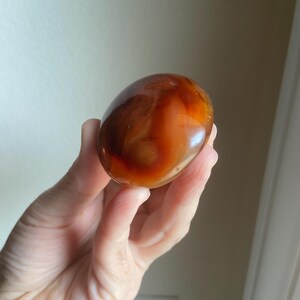 May include: A smooth, polished, oval-shaped carnelian stone featuring a translucent appearance with deep orange, reddish-brown, and amber color variations throughout its surface, displaying natural banding and a glossy, reflective finish.